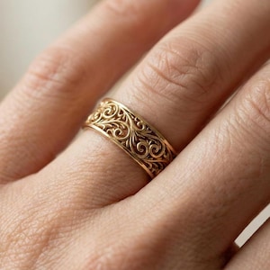 Può includere: Un anello color oro con un intricato design a filigrana a spirale. L'anello è indossato su un dito, mostrando il dettagliato lavoro a volute. La fascia è larga circa 1,3 cm e l'anello sembra essere in metallo.