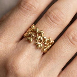 May include: A gold-colored ring featuring a repeating leaf design. The ring is worn on a finger, and the leaves are arranged in a continuous pattern around the band. The ring's design is intricate and detailed, with each leaf clearly defined.