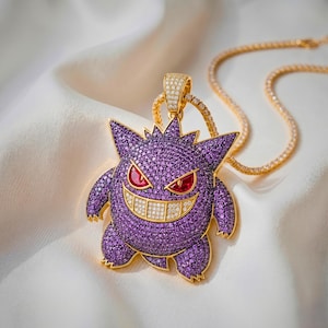 以下が含まれることがあります： 紫色のパヴェストーンで覆われたキャラクター型のチャームが付いたゴールドのペンダントネックレス。キャラクターは赤い宝石の目と、白いパヴェストーンで満たされた広い笑顔が特徴です。ペンダントは小さな透明な石が付いたゴールドのチェーンからぶら下がっています。