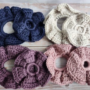 May include: A collection of handmade crochet scrunchies in various colors, including navy blue, beige, purple, and dusty rose. The scrunchies have a flower-like design and are arranged on a wooden surface.