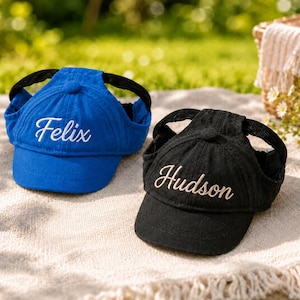 May include: A blue baseball cap with the name Felix embroidered in white and a black baseball cap with the name Hudson embroidered in white. Both caps feature ear holes on the sides and a button on the top, designed for pet headwear.