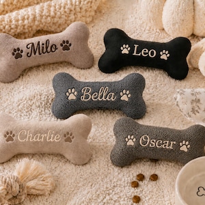 Op de afbeelding: Vijf botvormige pluche speeltjes van beige, zwarte en grijze stof. Elk speeltje is voorzien van geborduurde namen zoals Milo, Leo, Bella, Charlie en Oscar, geflankeerd door kleine pootafdrukpictogrammen. De speeltjes hebben een textuur oppervlak.