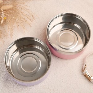 Puede incluir: Dos recipientes redondos de acero inoxidable para alimentos con exteriores rosa pastel y morado. Los recipientes est&aacute;n abiertos, revelando un interior brillante. Una correa blanca con herrajes dorados es visible.