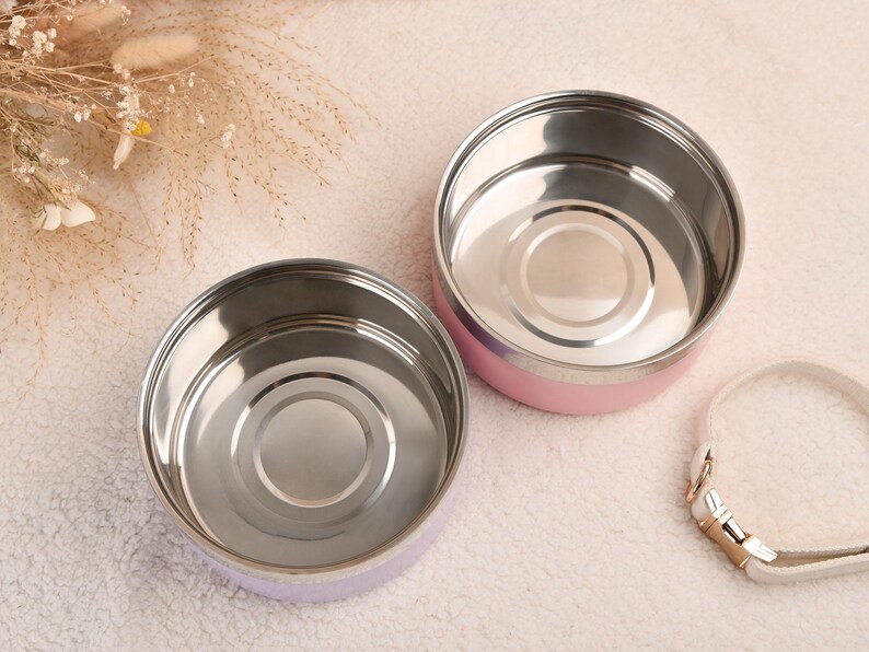 Puede incluir: Dos recipientes redondos de acero inoxidable para alimentos con exteriores rosa pastel y morado. Los recipientes est&aacute;n abiertos, revelando un interior brillante. Una correa blanca con herrajes dorados es visible.