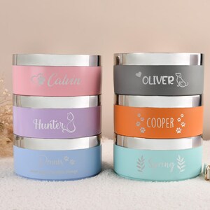 Puede incluir: Una pila de seis cuencos personalizados para mascotas en colores pastel: rosa, lila y azul a la izquierda, y gris, naranja y turquesa a la derecha. Cada cuenco tiene un borde plateado y un nombre grabado en la banda de color.