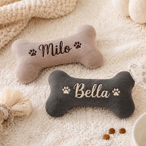 May include: Two bone-shaped pet toys made of textured fabric. The top toy is light beige with the name Milo embroidered in dark brown, flanked by two paw prints. The bottom toy is dark grey with the name Bella embroidered in white, also with two paw prints.