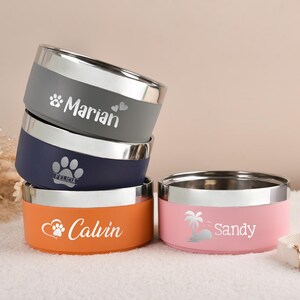 Puede incluir: Un conjunto de cuencos personalizados para mascotas en gris, azul marino y naranja, con un cuenco rosa. Cada cuenco tiene un borde plateado y un nombre impreso. Para mascotas.