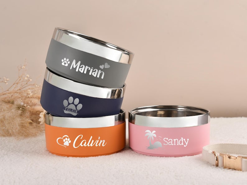 Puede incluir: Un conjunto de cuencos personalizados para mascotas en gris, azul marino y naranja, con un cuenco rosa. Cada cuenco tiene un borde plateado y un nombre impreso. Para mascotas.