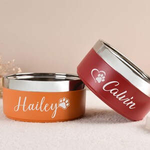 Puede incluir: Dos cuencos personalizados para mascotas. Uno es naranja con "Hailey" y una huella de pata, el otro es rojo con "Calvin", un coraz&oacute;n y una huella de pata. Ambos tienen un borde plateado.
