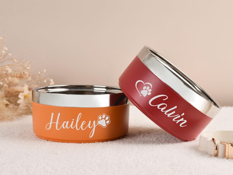 Puede incluir: Dos cuencos personalizados para mascotas. Uno es naranja con "Hailey" y una huella de pata, el otro es rojo con "Calvin", un coraz&oacute;n y una huella de pata. Ambos tienen un borde plateado.