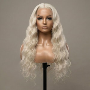 May include: A light blonde wig with long, wavy hair. The wig is styled on a mannequin head with a realistic face and makeup. The hair has a natural-looking hairline and is parted on the side.