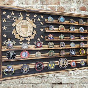 May include: A rectangular wooden display case featuring a charred finish and a black-painted flag design with stars. The case includes horizontal shelves holding various circular challenge coins and a prominent United States Coast Guard emblem.