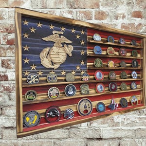 May include: Wooden display case shaped like an American flag, featuring a blue field with stars and red and white stripes. The case holds numerous military challenge coins. A wooden emblem of the Marine Corps is affixed to the blue field.