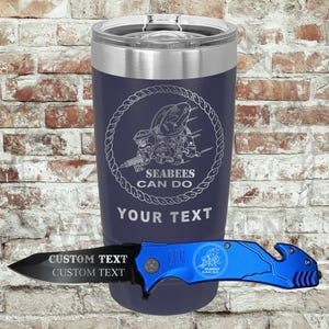 以下が含まれることがあります： シルバーの蓋が付いたネイビーブルーの断熱タンブラー。"SEABEES CAN DO"の文字とカスタムテキスト用のスペースがある円形デザイン。青と黒のユーティリティナイフがタンブラーの前に置かれています。