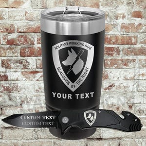 May include: Black tumbler with a silver rim and lid, featuring the text "MILITARY WORKING DOG" and a dog silhouette. Below the tumbler is a black pocket knife with the text "CUSTOM TEXT". The background is a brick wall.