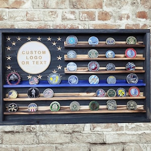 May include: A wooden display case in the shape of an American flag, with a dark blue frame and shelves. The case is filled with various challenge coins. A circular area is available for custom text or a logo.