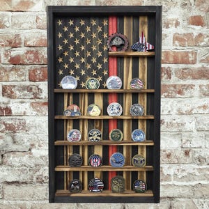 May include: A wooden display case with a rustic American flag design. The case features multiple shelves holding various challenge coins and decorative items. The flag design includes stars, stripes, and a red stripe, all within a black frame.