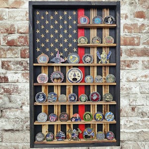 May include: A rectangular wooden display case featuring a dark-stained frame and a back panel painted with a stylized American flag design, including a vertical red stripe. The case has six horizontal shelves holding various circular metal coins.