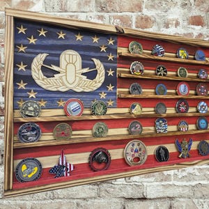 May include: Wooden coin display case shaped like an American flag, with red and white stripes and a blue field with stars. The case holds numerous military challenge coins. A wooden emblem is in the upper left corner.