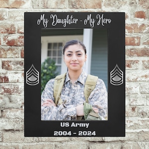 Puede incluir: Marco de fotos negro con el texto "My Daughter - My Hero" y "US Army 2004-2024". El marco presenta una foto de una mujer con uniforme militar. Insignias de rango militar plateadas a cada lado del marco.