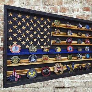 May include: A wooden display case shaped like an American flag, with a black frame and a blue stripe. The flag design features gold stars and wooden shelves holding various collectible coins. The background is a brick wall.