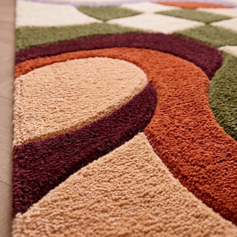 May include: A tufted rug featuring a geometric pattern with curved and checkered shapes in shades of olive green, deep burgundy, burnt orange, and cream. The surface has a thick, textured pile with distinct color-blocked sections throughout.