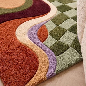 May include: A tufted area rug featuring a combination of patterns, including large flowing curves in shades of rust orange, deep burgundy, cream, and lavender, alongside a section of green and light green checkered square blocks.