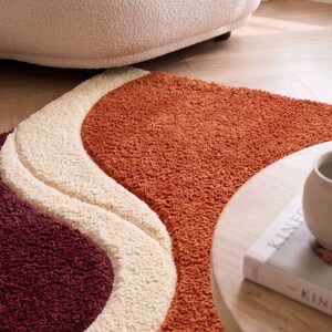 May include: A plush area rug featuring a wavy, abstract design with three distinct color sections. The rug displays a deep burgundy, a creamy off-white, and a vibrant burnt orange, all rendered in a thick, textured, high-pile shag material.