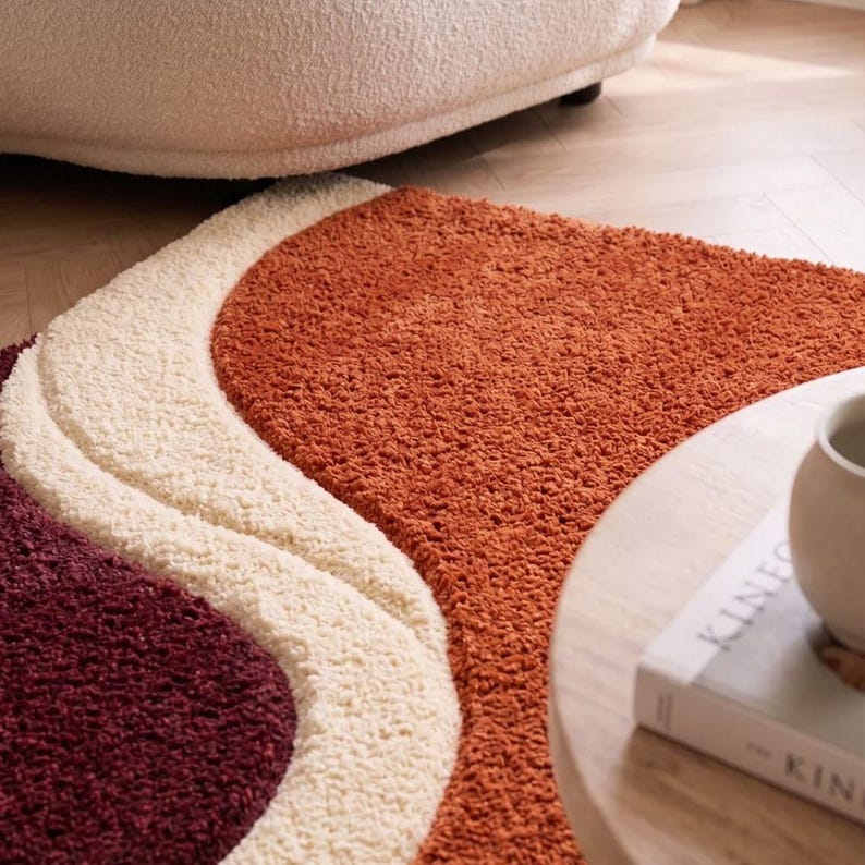 May include: A plush area rug featuring a wavy, abstract design with three distinct color sections. The rug displays a deep burgundy, a creamy off-white, and a vibrant burnt orange, all rendered in a thick, textured, high-pile shag material.