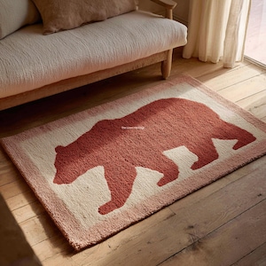 Può includere: Un tappeto rettangolare trapuntato con una silhouette di orso color terracotta centrata su uno sfondo color crema con un bordo color terracotta. Il tappeto ha una superficie testurizzata a riccioli e include il testo The Loom Heritage in piccolo.