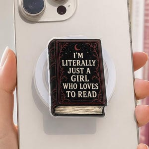 May include: A light-colored smartphone with a rectangular phone grip featuring a dark book design. The book cover has red filigree, a crescent moon, and the text: I'M LITERALLY JUST A GIRL WHO LOVES TO READ. The pages of the book are visible at the bottom.