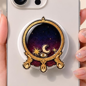 May include: A white smartphone with a circular phone grip attached to the back. The grip features a gold-toned frame surrounding a dark purple starry sky design, accented with a crescent moon, a star, and an eye symbol above red drapery.