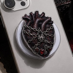 May include: A silver smartphone with a circular phone grip featuring a dark, anatomical heart wrapped in silver chains with a central padlock and small red heart accents. The phone grip is centered on the back of the device, which has a triple-lens camera.