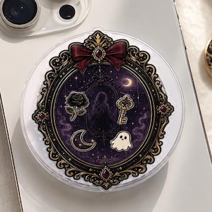 May include: A white smartphone with a circular phone grip featuring a dark purple ornate frame, a red bow, and illustrations of a black rose, a golden key, a crescent moon, and a small white ghost against a starry, dark background.