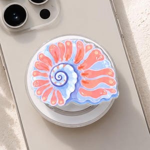 May include: A light-colored smartphone featuring a circular phone grip attached to the back. The grip displays a detailed illustration of a spiral seashell with vibrant coral-orange ridges and a soft periwinkle blue base, set against a white background.