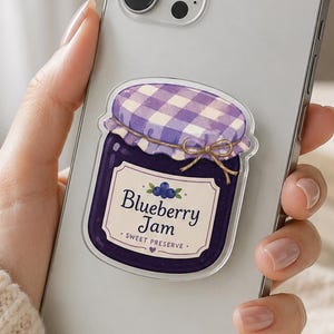 May include: A silver smartphone featuring a sticker of a dark purple jar of blueberry jam. The jar has a white label with the text Blueberry Jam and Sweet Preserve, topped with a purple and white checkered lid tied with a thin brown string.
