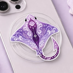 May include: A purple stingray-shaped phone grip featuring intricate white filigree patterns, small shell motifs, and star accents, attached to the back of a silver smartphone. The design includes a curved tail and symmetrical wing-like fins.
