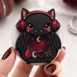 May include: A circular phone grip featuring a black bat character with red eyes and red headphones. The bat's body displays a dark red night sky scene with a crescent moon charm, a red droplet, a silver crescent moon, a music note, and a cassette tape.