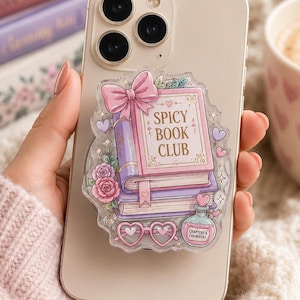 May include: A phone case with a clear pop-socket featuring a stack of books, roses, and the text "Spicy Book Club." The design includes a pink bow, heart-shaped glasses, and a potion bottle. The phone is held in a person's hand.