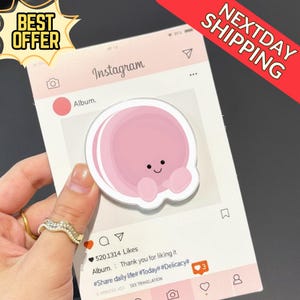 May include: A pink sticker with a cartoon character is held in front of a pink Instagram-themed background. The background includes the Instagram logo, a photo frame, and text that reads "Album." The sticker has a smiling face.