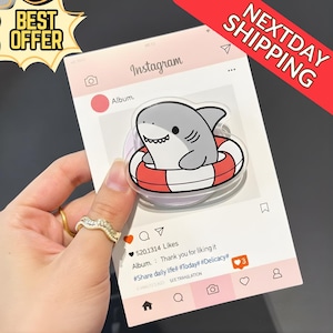 May include: A clear phone grip featuring a cartoon shark in a red and white life preserver. The grip is on a pink Instagram-themed card with the text "Album" and "520,1314 Likes". The card also has the text "BEST OFFER" and "NEXTDAY SHIPPING".