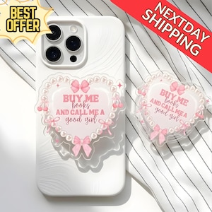 May include: White phone case with a heart-shaped pop socket that reads "BUY ME books AND CALL ME A good girl". The pop socket features pink bows and a pearl border. Another pop socket is visible.