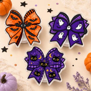 May include: Three decorative hair bows featuring Halloween themes. One orange bow displays bats and spiderwebs. A purple bow features white ghosts and spiderwebs. A second purple bow shows black cats with yellow eyes and stars.