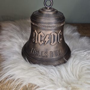 Op de afbeelding: Een bronzen bel met de tekst "AC/DC" en "HELLS BELL" in een vetgedrukte lettertype. De bel heeft een lus aan de bovenkant en staat op een witte, zachte ondergrond. De bel is een decoratief item.