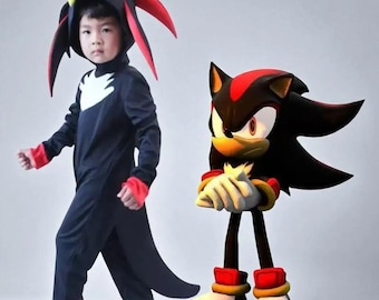 May include: A child in a black and red Shadow the Hedgehog costume, complete with a hood and gloves. A Shadow the Hedgehog figure stands nearby. The costume features a white chest emblem and red accents. The figure has red and white shoes.