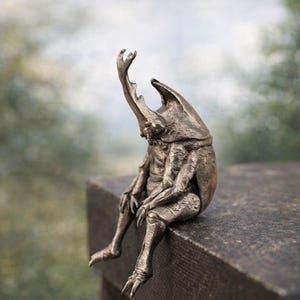 Puede incluir: Una escultura de bronce de una criatura parecida a un escarabajo sentada en una repisa. La escultura tiene detalles intrincados, incluyendo un cuerno prominente y extremidades articuladas. La criatura está representada con la cabeza baja y un brazo levantado, creando una pose pensativa.