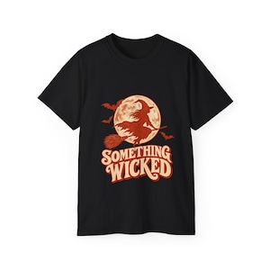Può includere: Una t-shirt nera a maniche corte con una stampa grafica di una strega che vola su una scopa davanti a una grande luna piena arancione con pipistrelli. Sotto la luna, il testo SOMETHING WICKED è stampato in un carattere arancione audace.