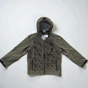 Könnte beinhalten: Olivgrüne Kapuzenjacke mit durchgehendem Reißverschluss und zwei Vordertaschen. Die Jacke hat ein sich wiederholendes Muster und ein Logo auf der Brust. Die Ärmel haben schwarze Verzierungen und ein Etikett ist angebracht.