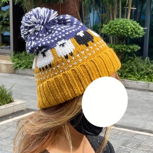 May include: A knit beanie featuring a mustard yellow ribbed cuff and a pattern of white and black sheep across the center. The top section is navy blue with a white geometric design, finished with a large navy and white pom-pom.