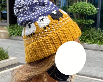 Può includere: Un berretto in maglia con bordo a coste giallo senape e un motivo di pecore bianche e nere al centro. La parte superiore è blu navy con un disegno geometrico bianco, completata da un grande pon pon blu navy e bianco.